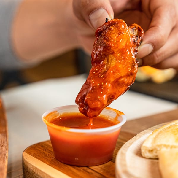 A hand dips a saucy chicken wing into a small cup of red hot sauce.