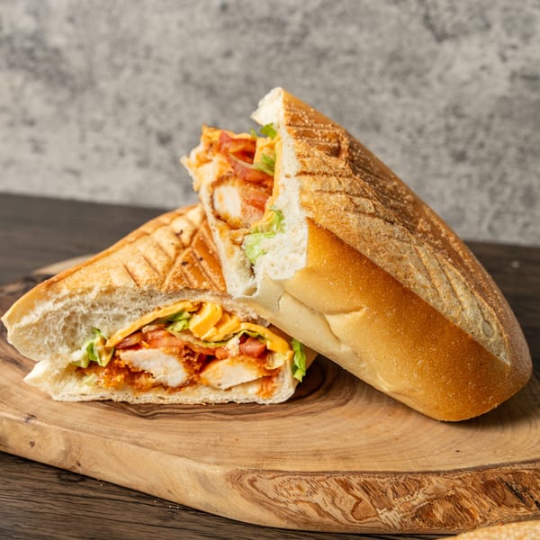 A grilled chicken sandwich with cheese, lettuce, and tomatoes, cut in half on a wooden board.