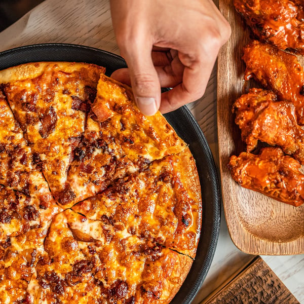 A hand grabbing a slice of Singas Famous Pizza next to a wooden tray of buffalo wings.