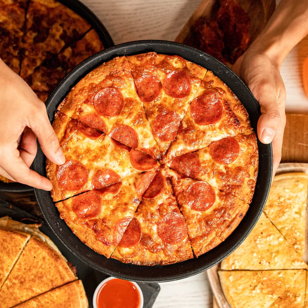 Hands holding a freshly baked, sliced pepperoni pizza in a black pan, with other pizzas in the background.