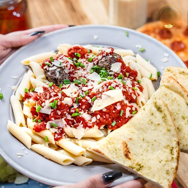Penne pasta with meatballs, red sauce, parmesan, chives, and flatbread, held by hands.