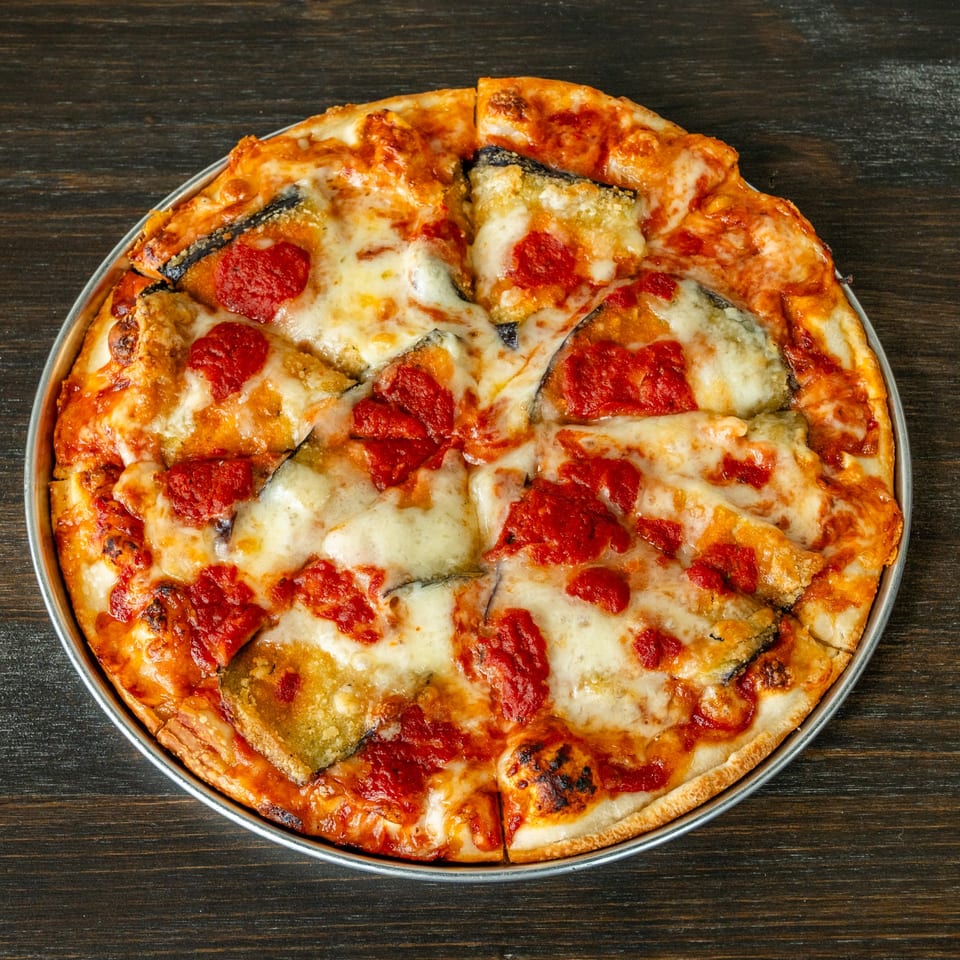 A freshly baked eggplant parmesan pizza, cut into slices, in a metal pan on a dark wooden table.