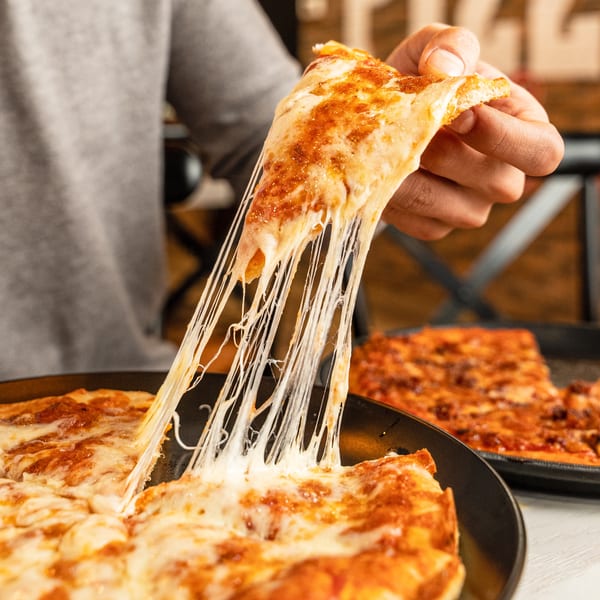 A hand lifts a slice of Singas Famous Pizza, stretching melted cheese from the pan.