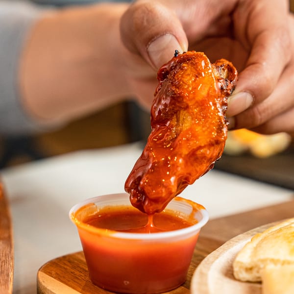 A hand dips a saucy chicken wing into a small cup of red dipping sauce.