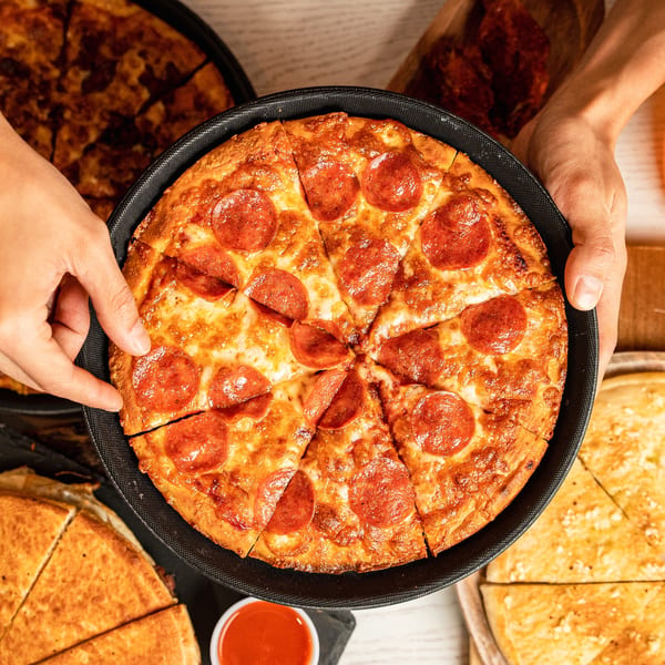 Hands holding a hot, sliced pepperoni pizza in a black pan, surrounded by other pizzas.