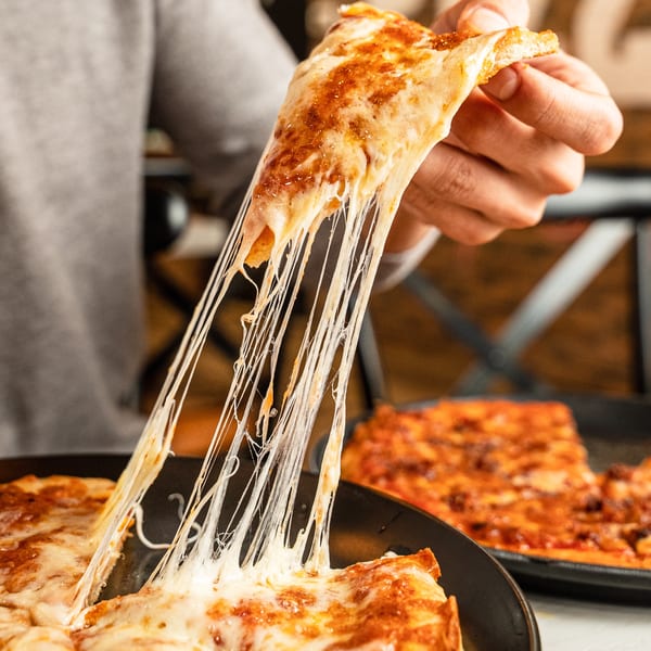 A hand lifts a slice of Singas Famous Pizza, with long, stretchy cheese pulls.
