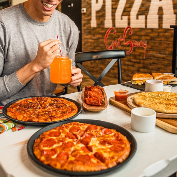 A person smiling, drinking orange juice, with two pizzas, wings, and garlic bread on a table.
