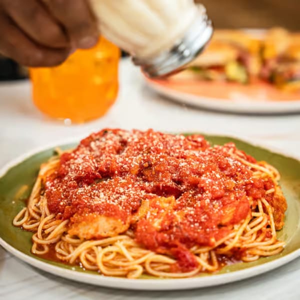 A person sprinkles parmesan cheese over a plate of spaghetti with red sauce and chicken.