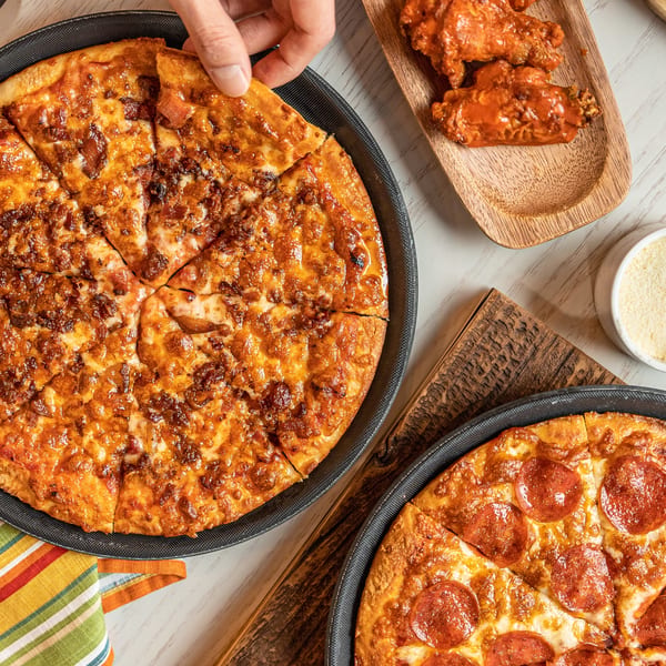 Hand grabbing a slice of bacon pizza, with a pepperoni pizza and buffalo wings nearby.