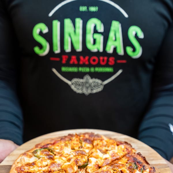 A person wearing a Singas Famous Pizza shirt holds a freshly baked pizza on a wooden peel.