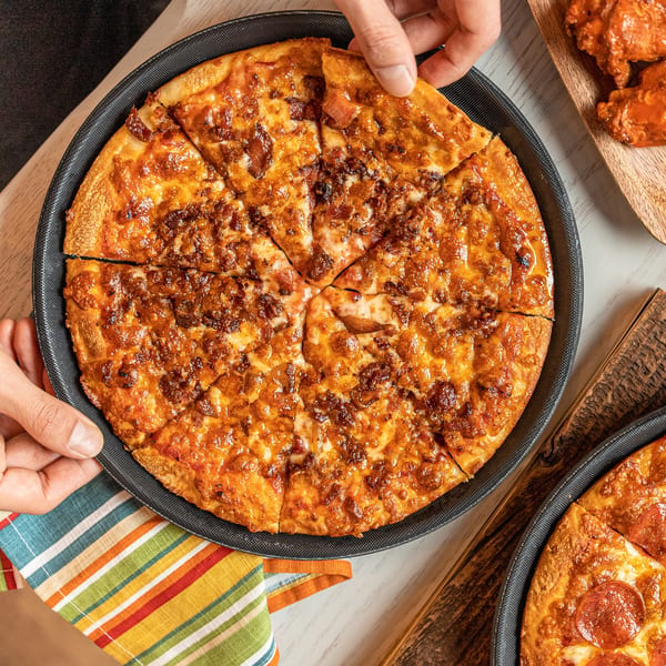 Hands taking a slice of Singas Famous Pizza with bacon topping, with another pepperoni pizza nearby.