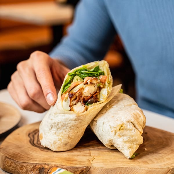 A person's hand reaching for a chicken and lettuce wrap on a wooden board.