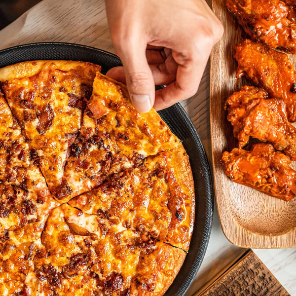 A hand grabs a slice of Singas Famous Pizza next to a tray of buffalo wings.