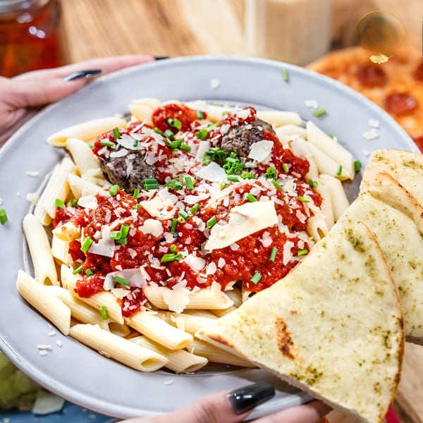Penne pasta with meatballs, red sauce, parmesan, chives, and garlic bread on a gray plate.