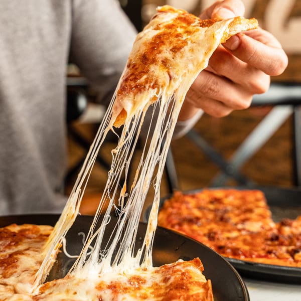A hand lifts a slice of Singas Famous Pizza, with long, stretchy cheese pulls.