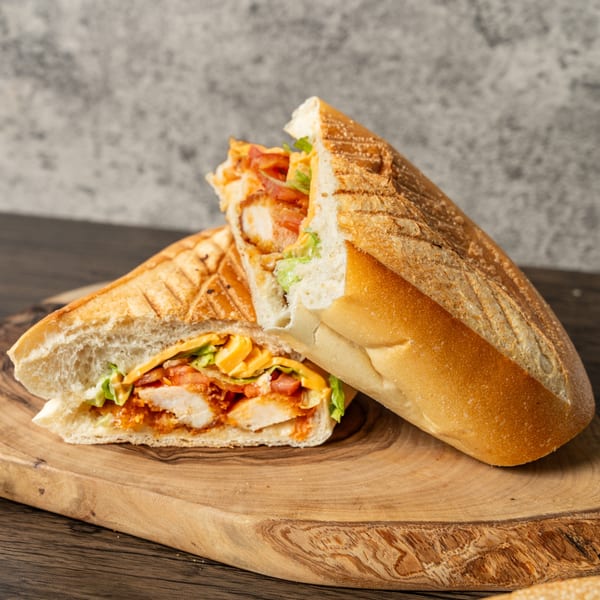 A grilled chicken sandwich with lettuce, tomatoes, and sauce, cut in half on a wooden board.