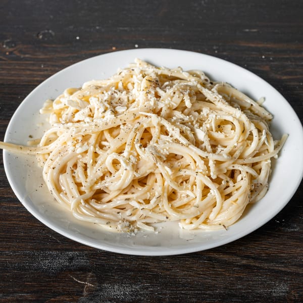 A white plate of spaghetti with creamy sauce, crumbled cheese, and herbs on a dark wooden table.