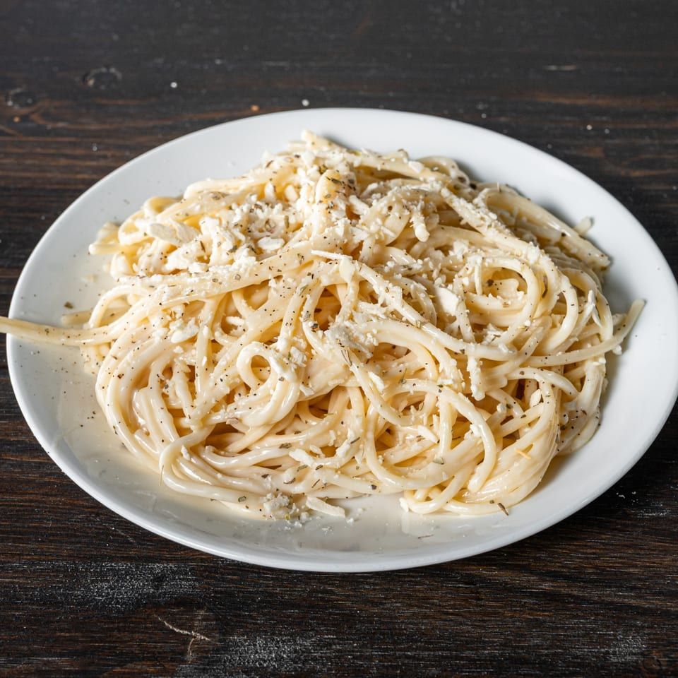 A white plate of spaghetti with creamy sauce, crumbled cheese, and herbs on a dark wooden table.