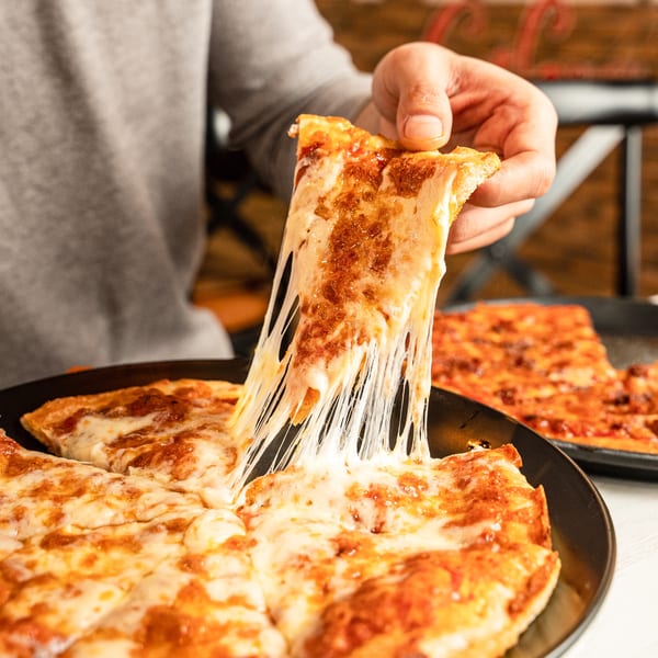 A hand pulls a slice of Singas Famous Pizza, stretching the melted cheese, with another pizza in the background.