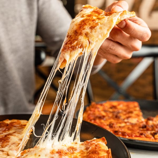 A hand lifts a slice of Singas Famous Pizza, stretching melted cheese into long strands.