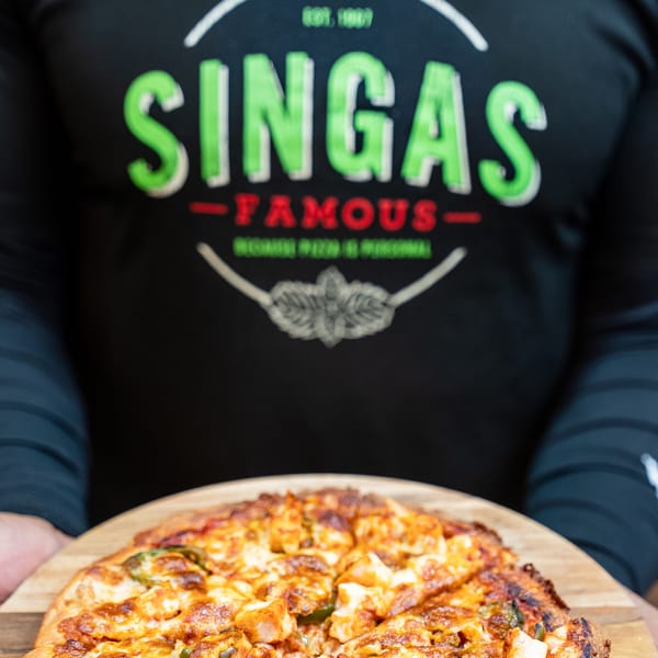 A person in a Singas Famous Pizza shirt holds a freshly baked pizza with chicken and peppers.