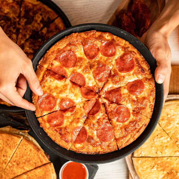 Hands holding a freshly baked pepperoni pizza in a pan, surrounded by other pizzas and dipping sauce.