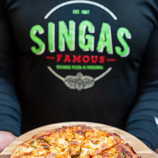 A person wearing a Singas Famous Pizza shirt holds a freshly baked pizza on a wooden peel.