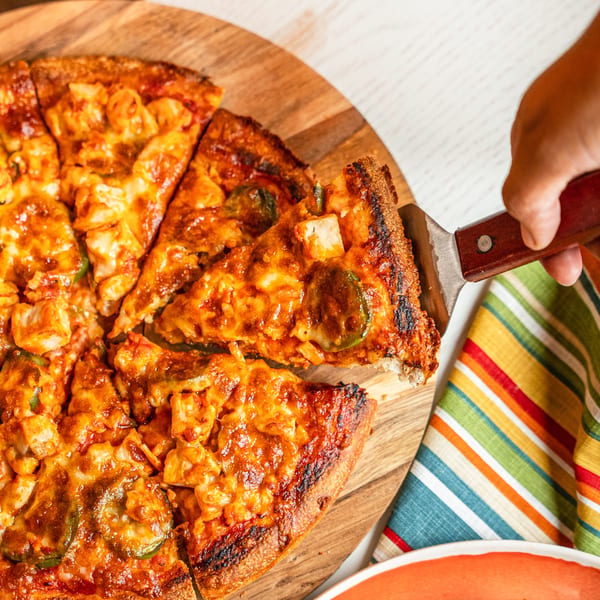 A hand uses a spatula to lift a slice of Singas Famous Pizza with chicken and jalapeños.